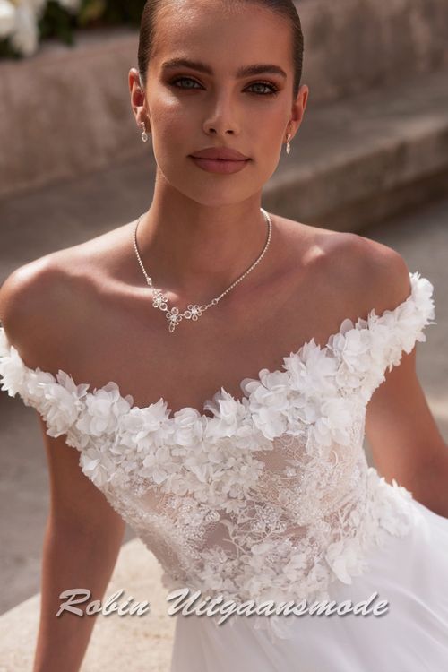 Close-up van de Off-shoulder trouwjurk met kanten lijfje,  een bloemen versierde halslijn, sluikvallende chiffon rok met semi lange sleep